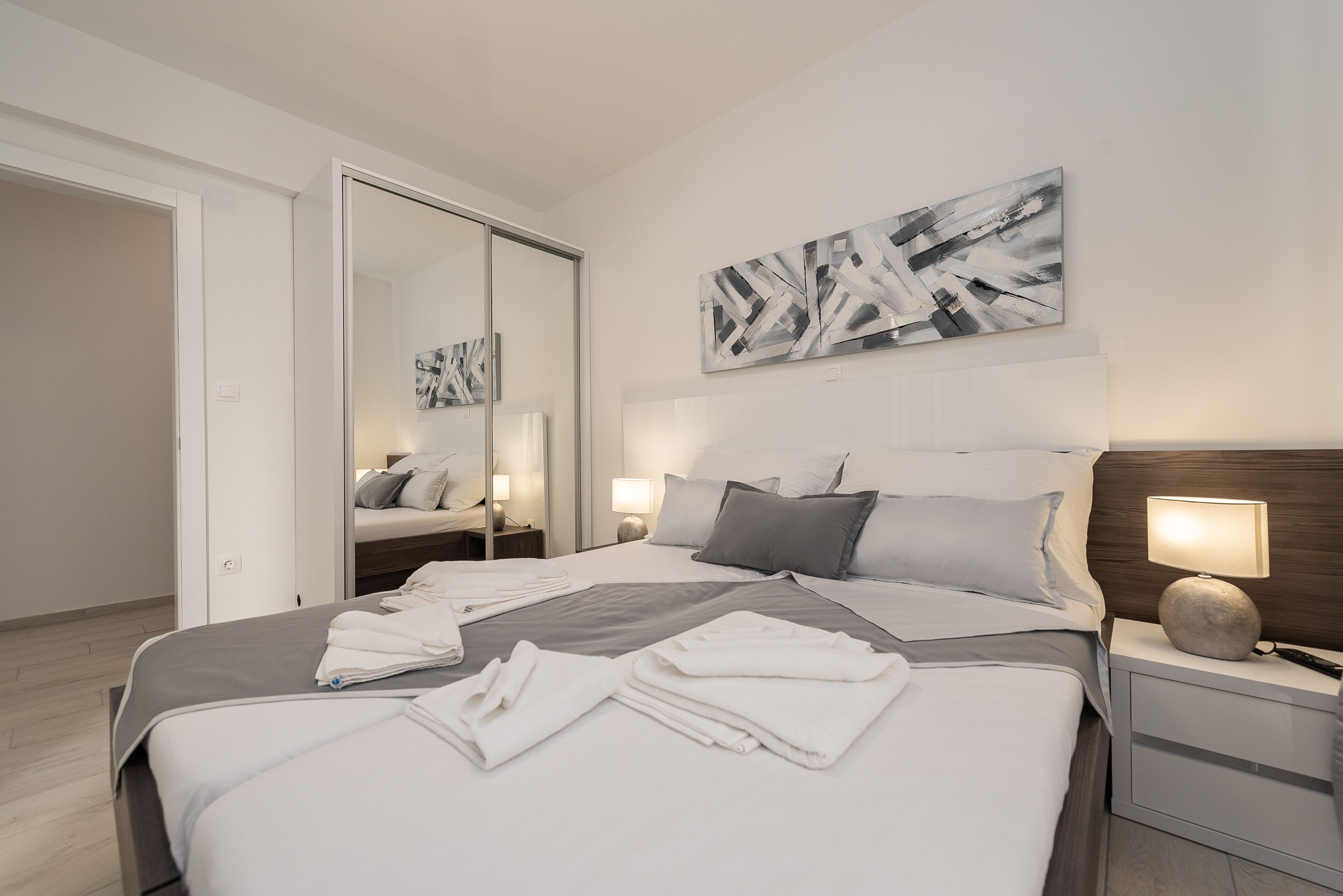 A modern hotel room with a large bed, a bedside table with a lamp, a mirror, and abstract wall art.