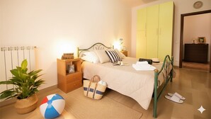 A bedroom with a bed, bedside tables, a nightstand, a lamp, a potted plant, a beach ball, and a suitcase. - Appartamento Kennedy 1 (Diano Marina)