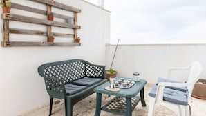En balkon med en træhylde med krukker, en grøn stol med hynder, en hvid stol og et lille bord med en bakke.
