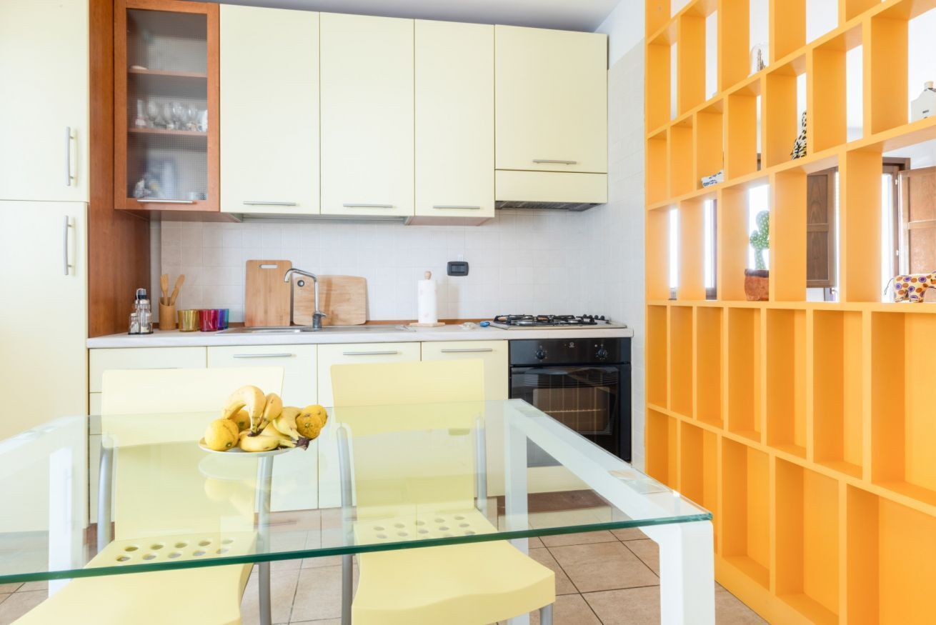 Une cuisine moderne avec des meubles blancs, une table à manger en verre et une étagère de couleur jaune orangé.