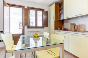 Una cocina moderna con una mesa de vidrio, sillas amarillas, armarios de madera y una ventana con contraventanas abiertas.