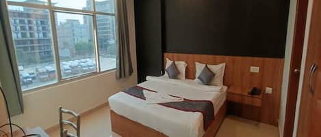 Habitación de hotel con una cama grande, un escritorio y una silla. Cuenta con un ventilador de techo, una ventana con vista a edificios y una puerta.