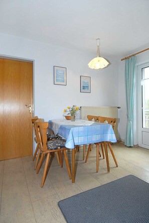 Un coin repas avec une table dressée pour quatre personnes, des chaises en bois, une nappe bleue, deux tableaux encadrés au mur, un luminaire suspendu et une porte avec une poignée en laiton.
