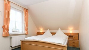 A bedroom with a wooden bed, two bedside tables with lamps, a window with curtains, and a radiator. - Vacation apartments in Huus Janssand in Bensersiel - Holiday apartment 1 (Esens)