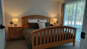 Un dormitorio con una cama de madera, dos mesitas de noche con lámparas, un atril y suelo alfombrado, además de una ventana con cortinas.