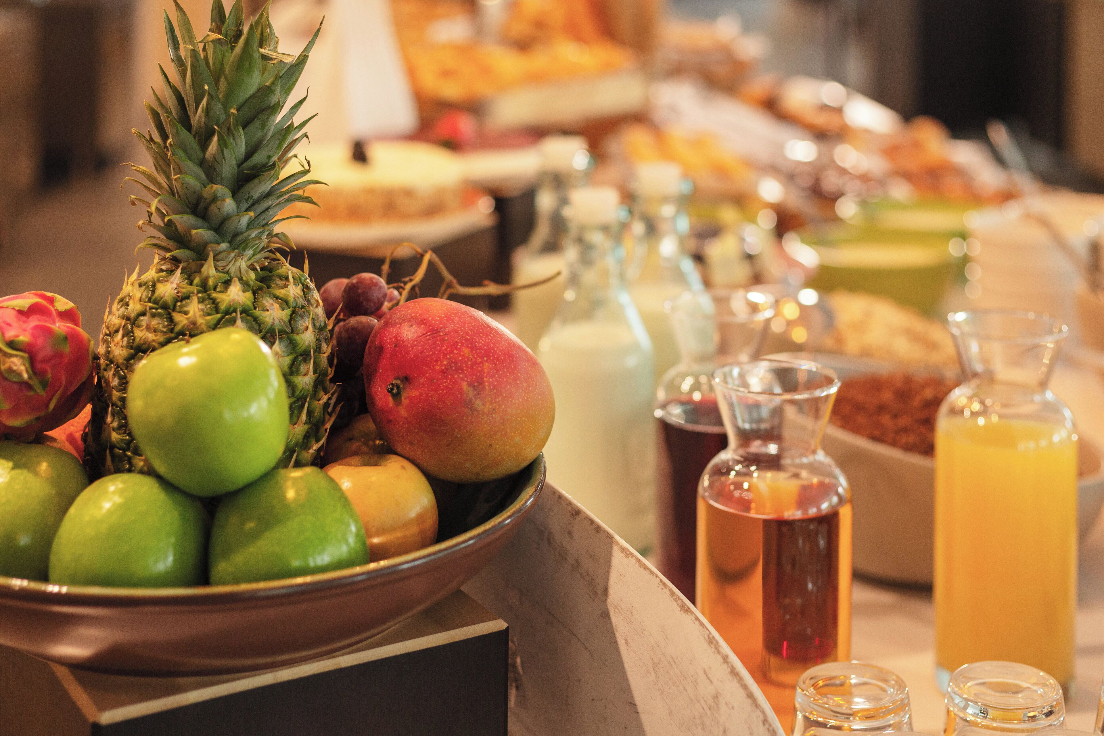 A fruit bowl with pineapple, mango, and apples, and several glass bottles with drinks.