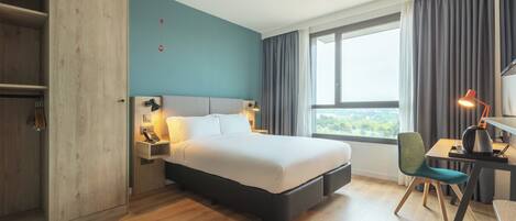 A modern hotel room with a large bed, a desk with a chair, a wardrobe, and a view of the outdoors through the window.