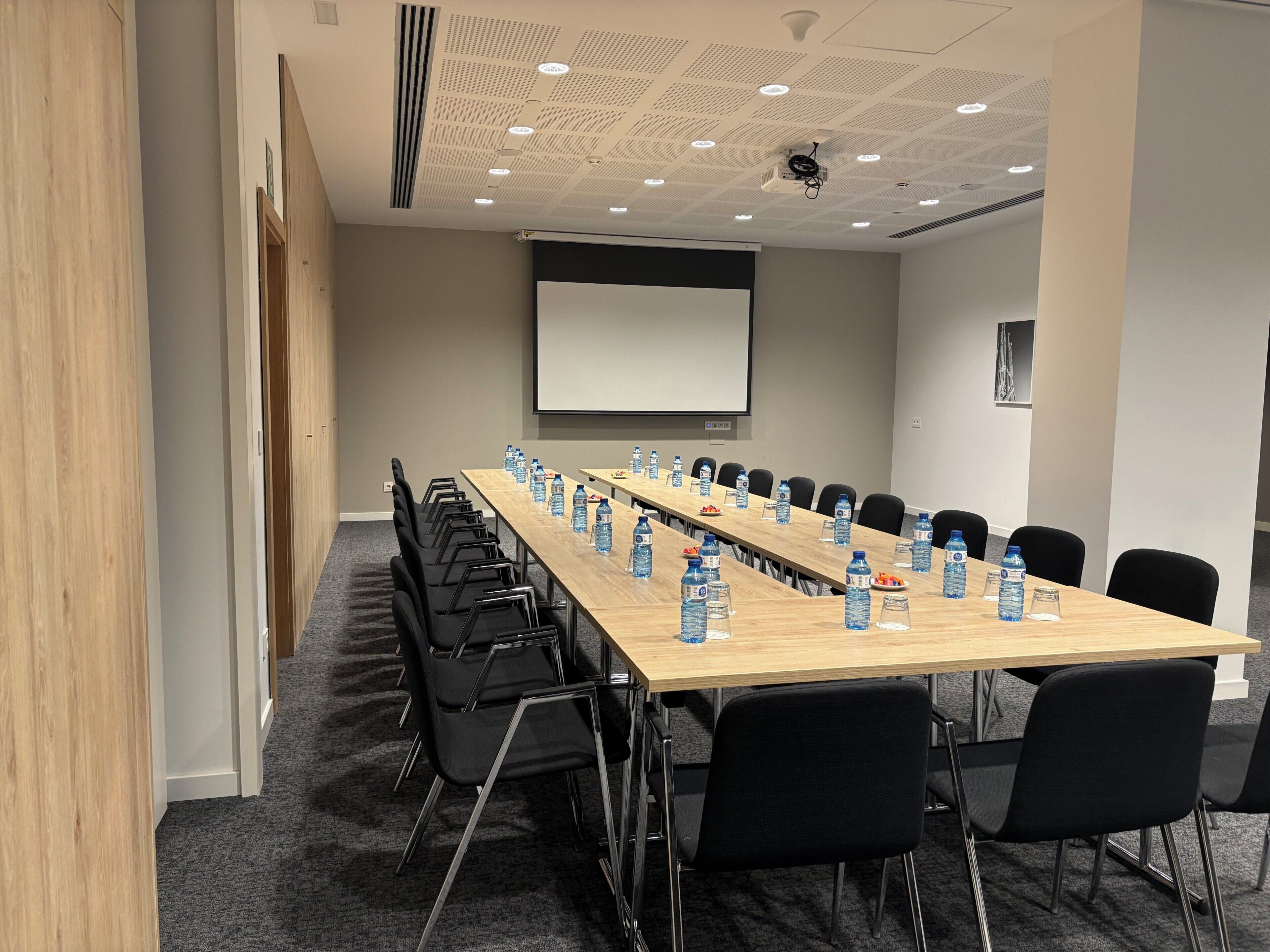 Un salón de conferencias con una larga mesa, sillas negras, botellas de agua y una pantalla de proyector.