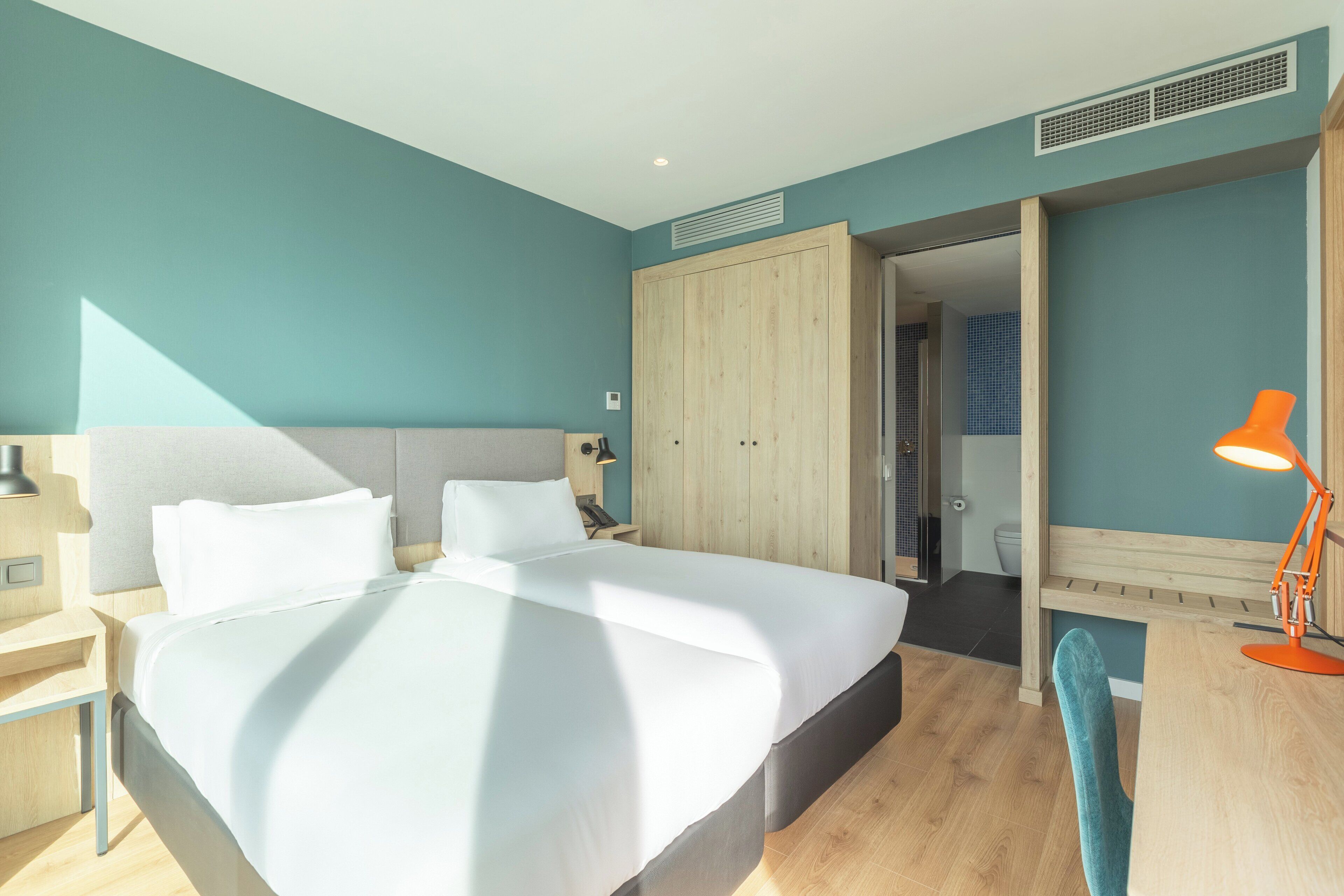 Una habitación de hotel moderna con una cama grande, un armario de madera, un escritorio con una silla y un baño visible a través de una puerta abierta.