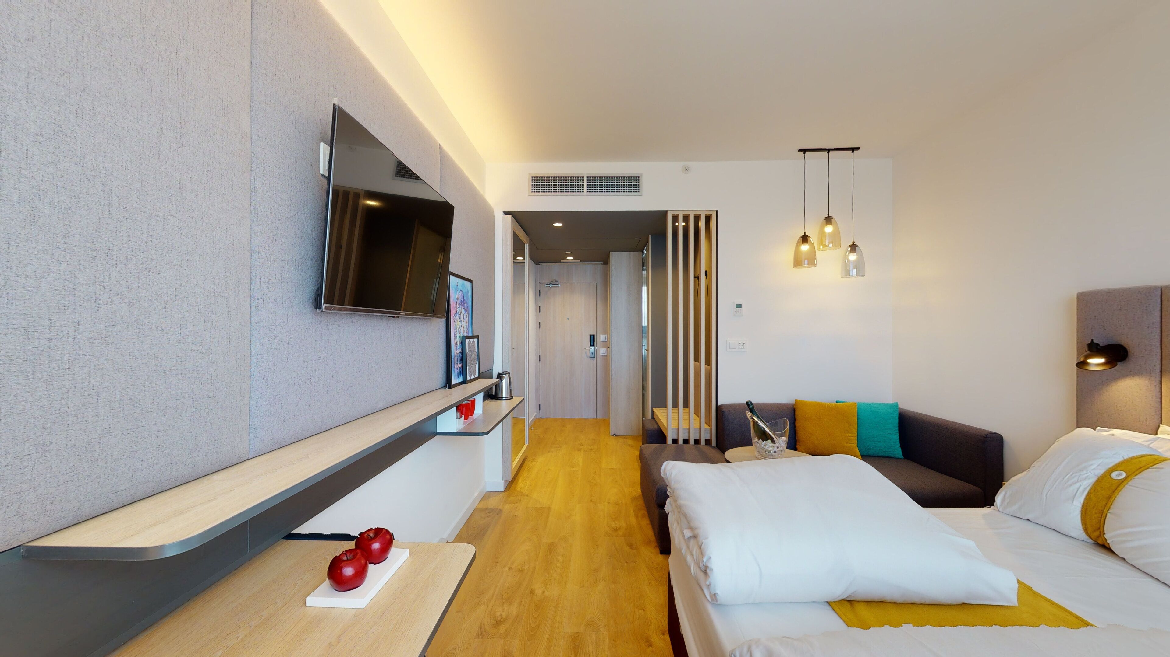 A modern hotel room with a wooden floor, a bed with white and yellow bedding, a sofa with colorful cushions, a mounted TV, and a small table with apples.