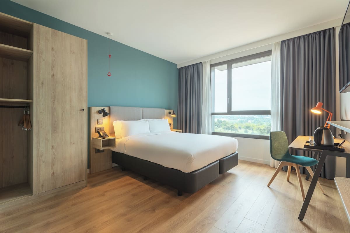 Une chambre d’hôtel moderne, dotée d’un grand lit, d’un bureau avec une chaise, d’une armoire et offrant une vue sur l’extérieur par la fenêtre.