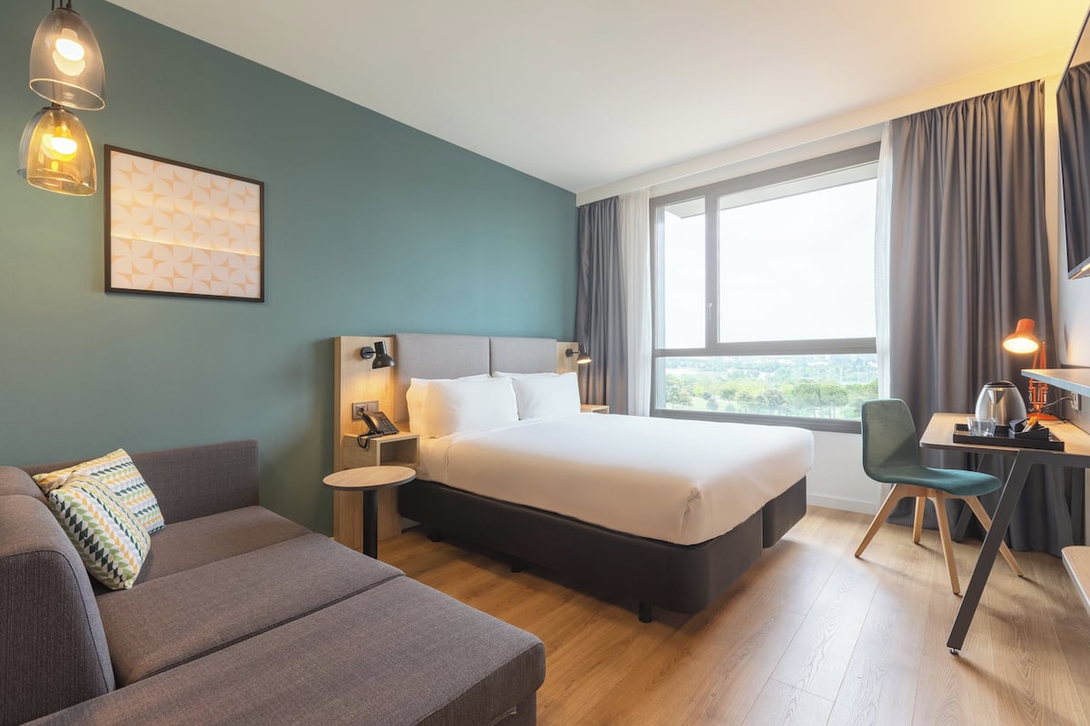 Une chambre d’hôtel comprenant un lit, un canapé, un bureau avec une chaise, une lampe et une fenêtre avec des rideaux.