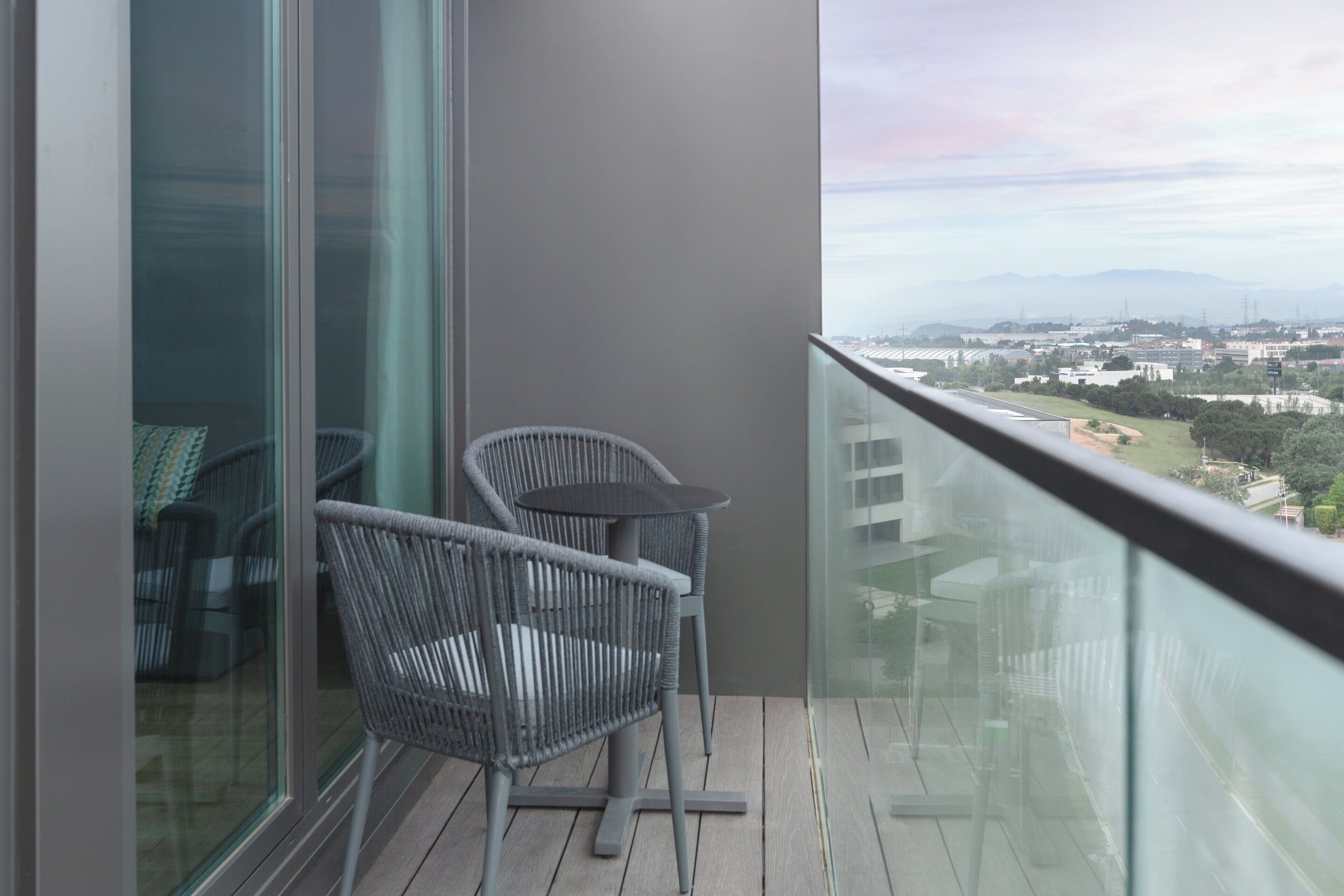 A balcony with wicker chairs and a table, overlooking a cityscape with buildings and a bridge.