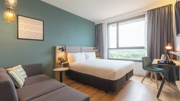 Habitación de hotel con cama, sofá, escritorio con silla, lámpara y ventana con cortinas.