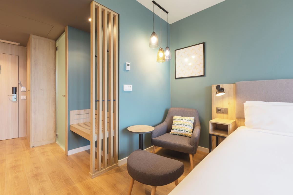 Une chambre d’hôtel moderne comprenant un lit, un coin salon avec un fauteuil et un pouf, une petite table, une lampe fixée au mur et un tableau encadré.