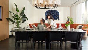 Een moderne eethoek met een grote tafel, zwarte stoelen en een kroonluchter. Achterin staat een bank met kussens en een potplant.