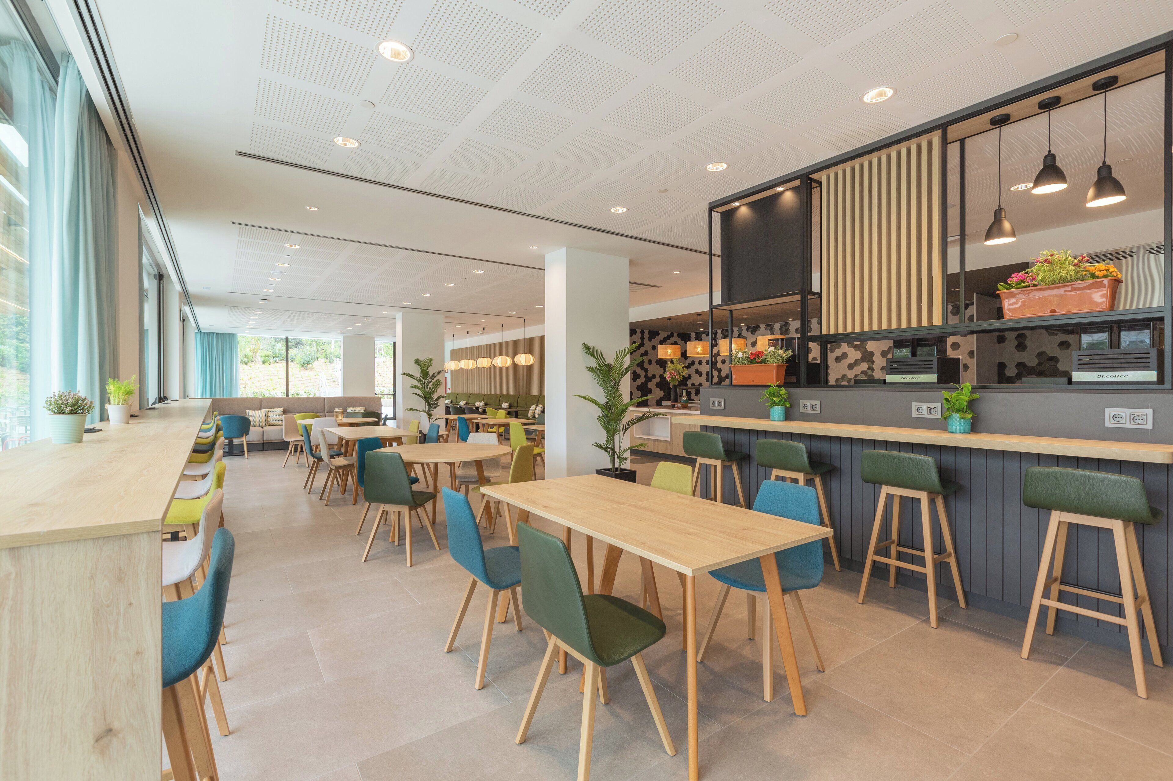 A modern restaurant with wooden tables and green chairs, a bar area with stools, and potted plants.