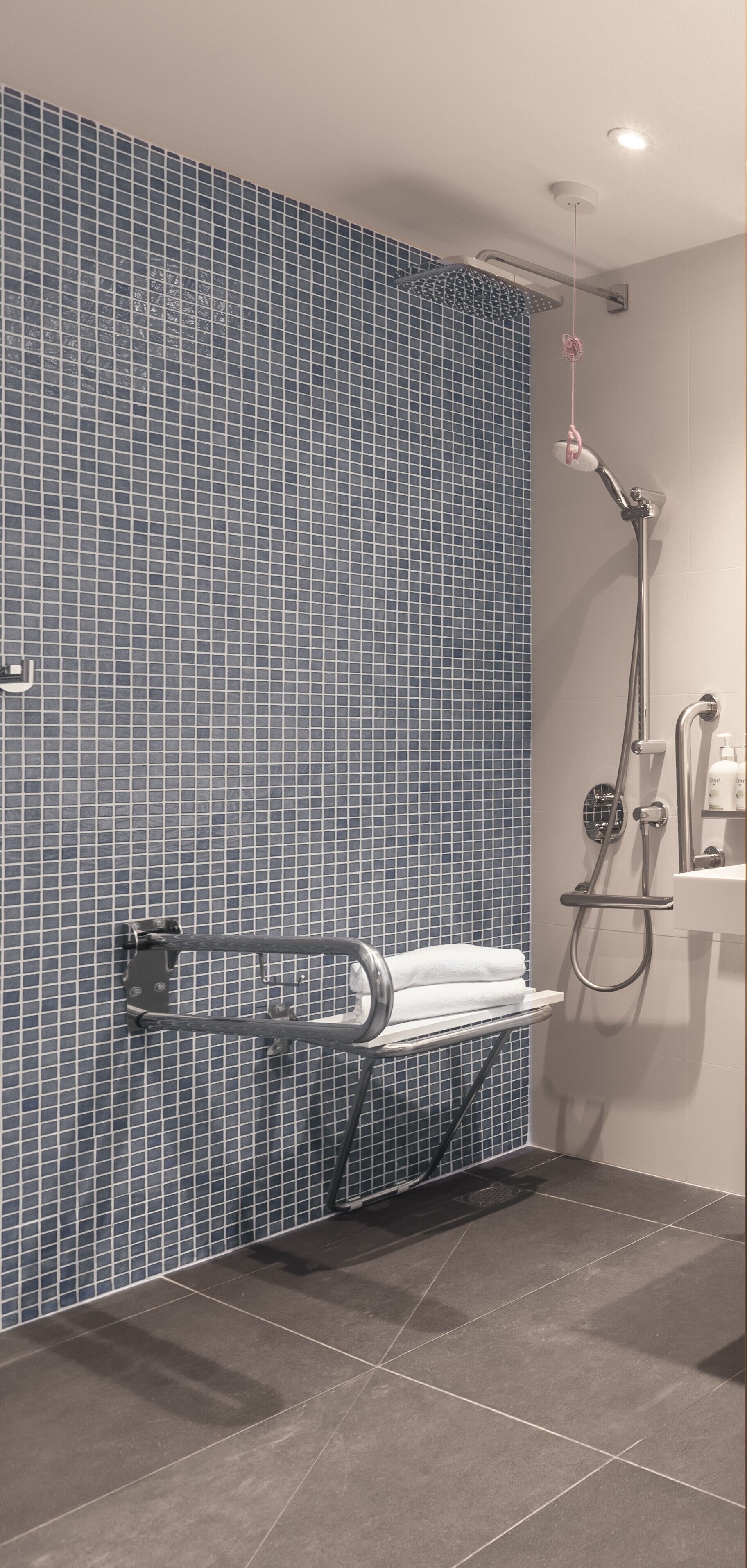 Un baño con pared de azulejos azules, ducha con cabezal de mano y un perchero con una toalla blanca.