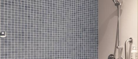 Un baño con pared de azulejos azules, ducha con cabezal de mano y un perchero con una toalla blanca.