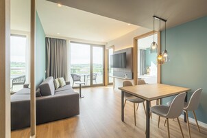 A modern living room with a sofa, dining table, chairs, a flat-screen TV, and a balcony with outdoor seating.