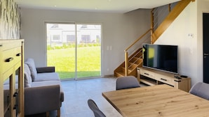 Un salon moderne avec une table à manger en bois, des canapés gris, un téléviseur et un escalier menant à l’étage supérieur.
