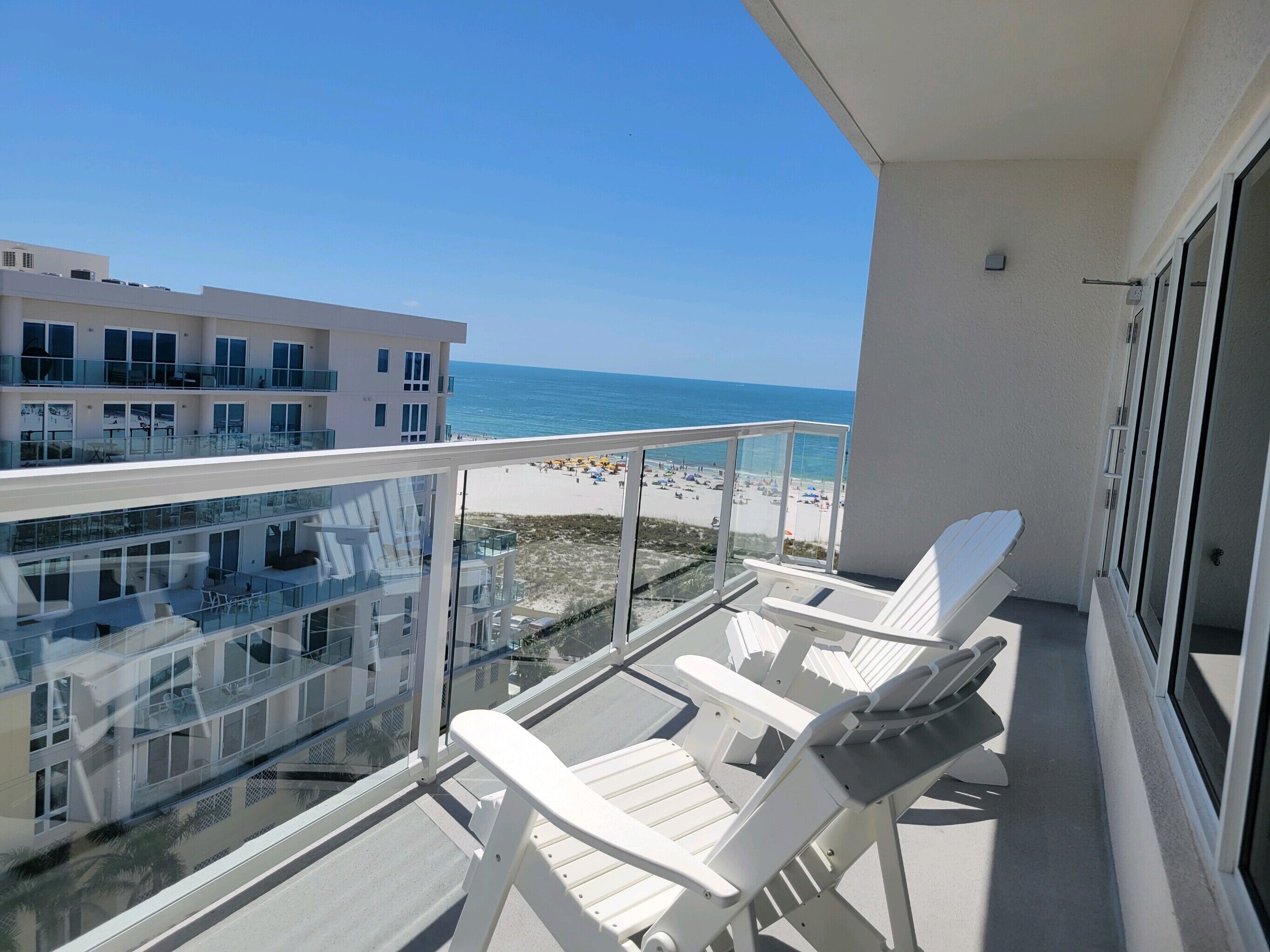 A balcony with white chairs and a glass railing overlooking a beach and a building.