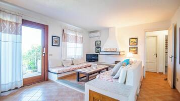 Uma sala de estar com um sofá, uma mesa de centro e uma televisão. Uma porta deslizante dá acesso a uma varanda.
