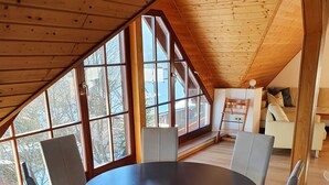 Amplia habitación con techo alto y ventanas grandes, con una mesa redonda y sillas, y vista al exterior.