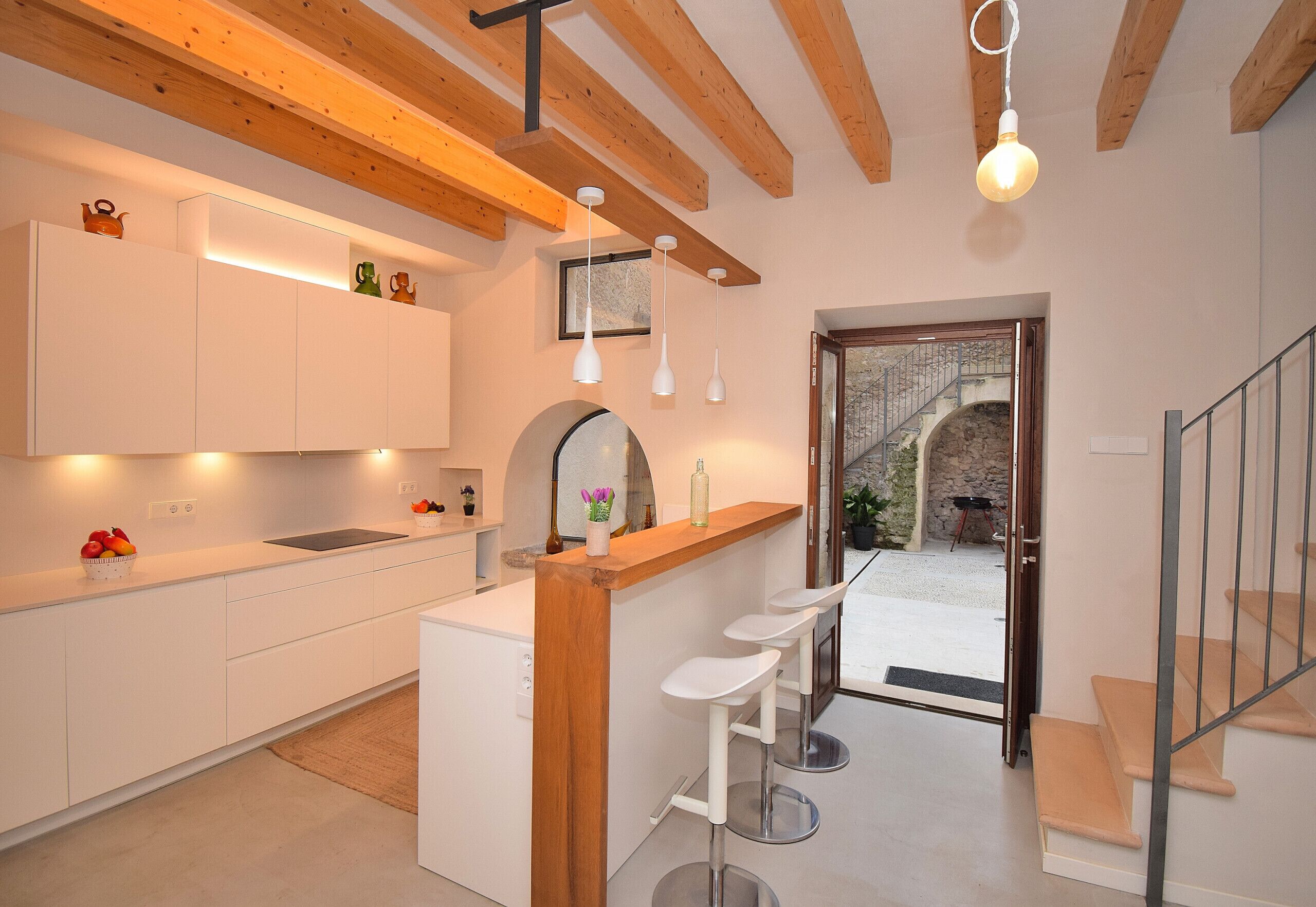 A modern kitchen with white cabinets, a wooden island, and bar stools. There is a staircase leading to a balcony with a stone wall.
