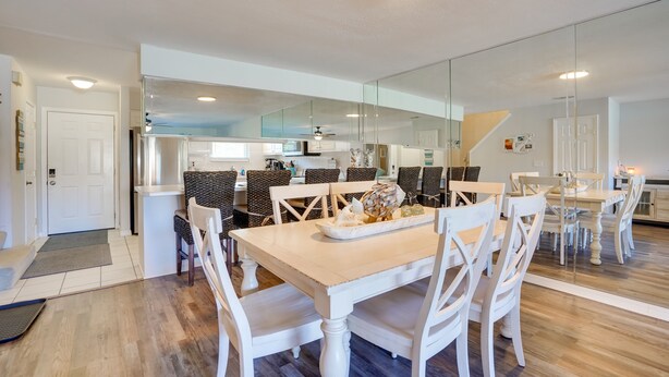 A modern kitchen with white cabinetry, a central island, and a dining area with white chairs and a wooden table. - Sun-Dappled PCB Retreat w/ Multiple Pools! (Panama City Beach)