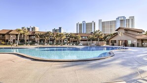 A swimming pool area with lounge chairs, palm trees, and a building with multiple floors in the background. - Sun-Dappled PCB Retreat w/ Multiple Pools! (Panama City Beach)