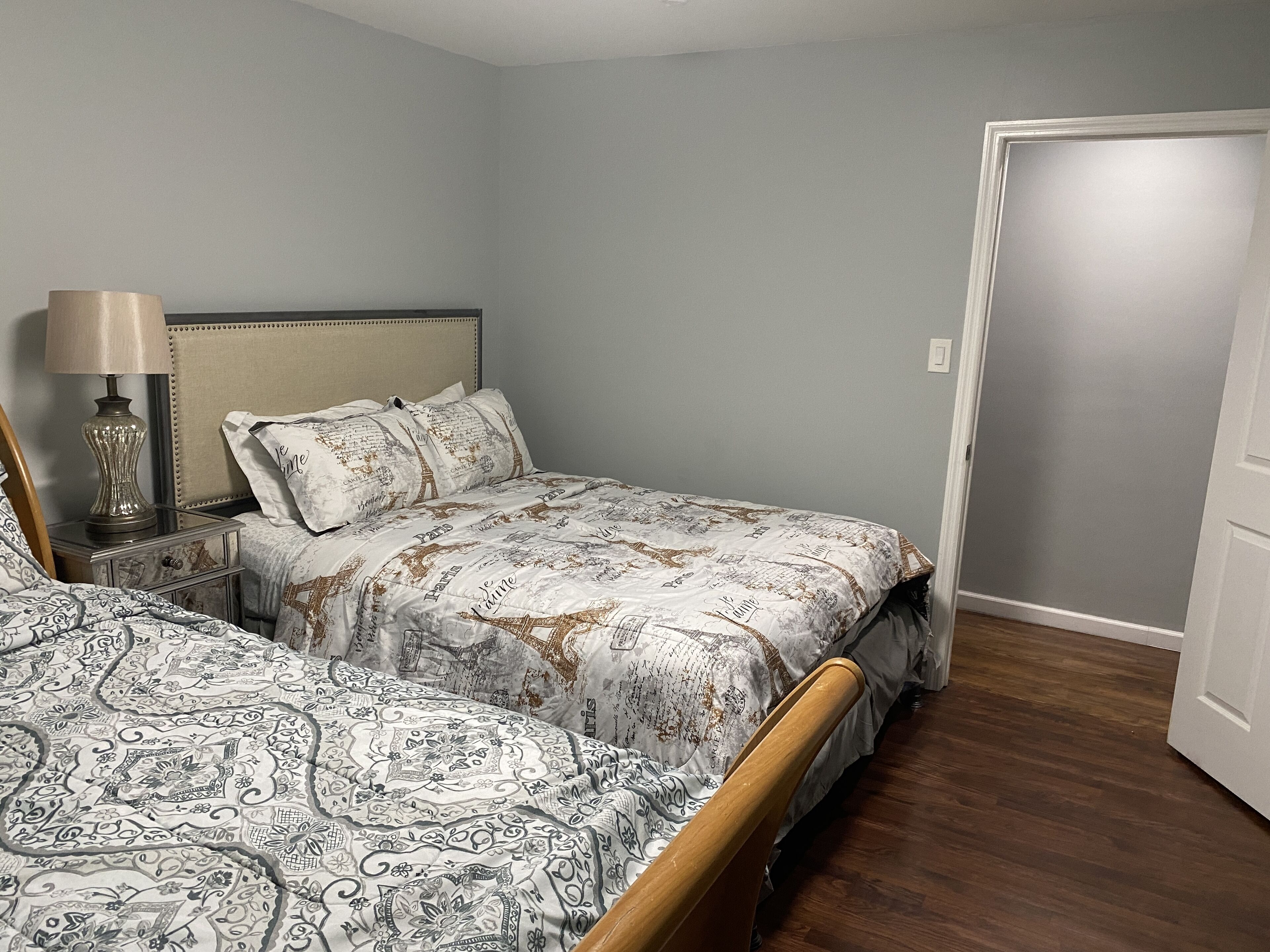 A bedroom with two beds, a nightstand with a lamp, and a door to another room.