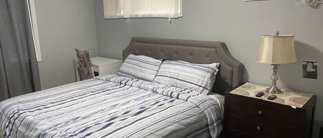 Un dormitorio con cama, mesita de noche, lámpara, aire acondicionado y ventana con cortinas.