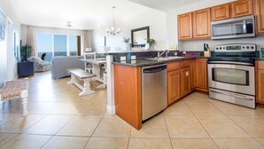 A modern kitchen with a central island, stainless steel appliances, wooden cabinets, and a dining area with a view. - Ariel Dunes I 1407 *Beach Access*3 pools* (Miramar Beach)