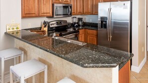 A kitchen with granite countertops, stainless steel appliances, wooden cabinets, and white bar stools. - Ariel Dunes I 1407 *Beach Access*3 pools* (Miramar Beach)