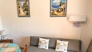A room with a grey sofa, two wall-mounted paintings, a small table with a lamp, and a bed with a checkered bedspread. - L'Amirauté by Interhome (Bormes-les-Mimosas)