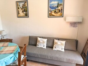 A room with a grey sofa, two wall-mounted paintings, a small table with a lamp, and a bed with a checkered bedspread.