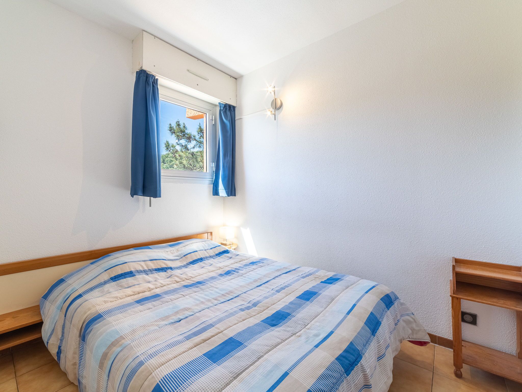 Un dormitorio con cama, ventana con cortinas, luz empotrada en la pared y mesita de noche de madera.