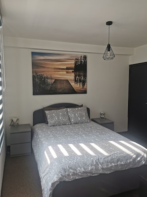 A bedroom with a bed, a nightstand, a lamp, and a large painting of a sunset over a lake. - Impasse 2. (Fosses-la-Ville)
