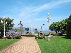 A paved pathway leading to a fountain with water jets, surrounded by lush greenery and flowering plants.