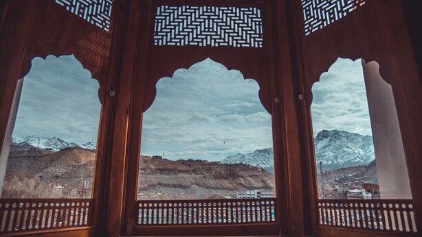 Una habitación con tres ventanas de madera con elaborados diseños de celosía, que ofrece vistas a montañas nevadas y un valle.