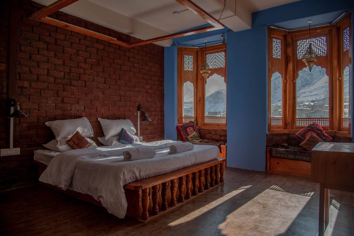 A bedroom with a brick wall, a wooden bed, a bench, and a view of mountains through a window with decorative wooden lattice.