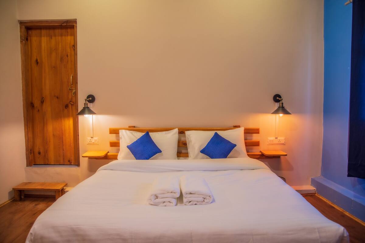 A neatly made bed with white linens and blue pillows, two bedside lamps, a wooden door, and a small wooden bench.