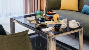 A modern living room with a glass coffee table, a sofa with cushions, and a dining area with a tray of snacks and drinks.