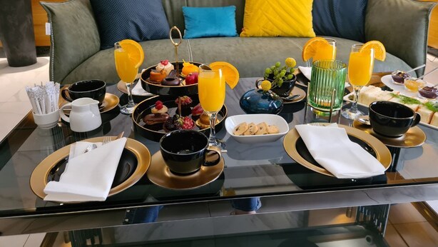 A breakfast buffet on a glass table with various dishes, drinks, and a sofa in the background.