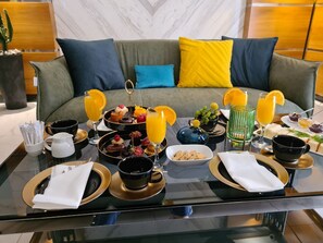 A breakfast buffet on a glass table with various dishes, drinks, and a sofa in the background. - ATECA HOTEL SUITES (Tashkent)