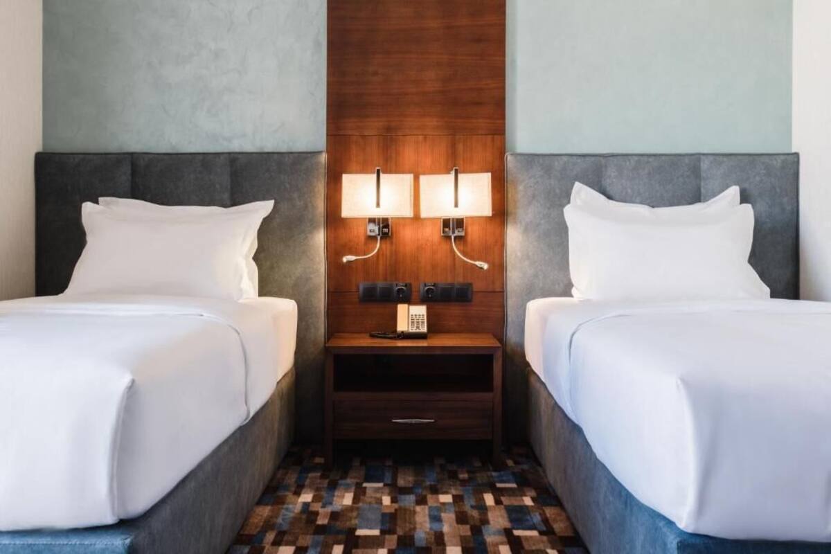 A hotel room with two beds, a wooden headboard, a nightstand with lamps, and a check-in counter.