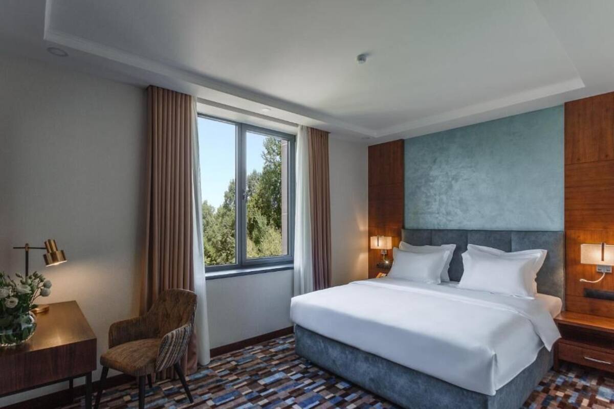 A hotel room with a large bed, a desk with a lamp, a chair, and a view of trees through the window.