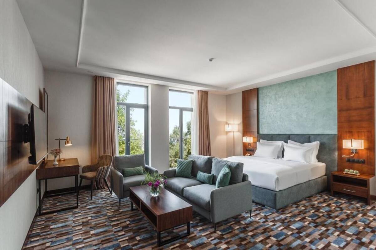 A hotel room with a large bed, a sofa, a coffee table, and a TV. There are windows with curtains, a lamp, and a wall-mounted TV.