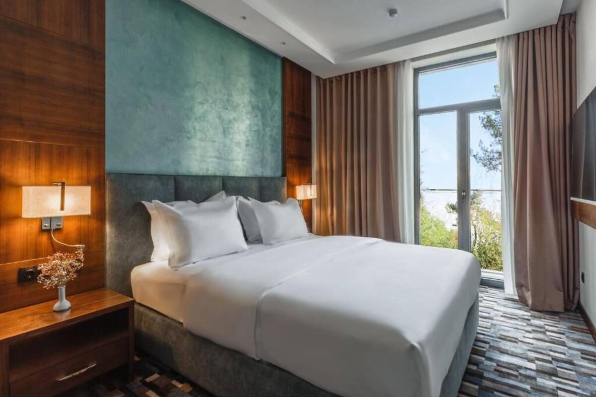 A hotel room with a large bed, two bedside lamps, a wooden nightstand, a vase of flowers, and a view of the outdoors through a large window.